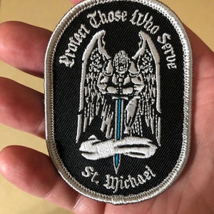 May include: A black embroidered patch with a white angel holding a sword. The text around the angel reads "Protect Those Who Serve" and "St. Michael".