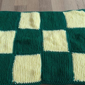 Könnte beinhalten: Eine handgestrickte Decke mit Schachbrettmuster. Die Decke hat abwechselnd Quadrate in Dunkelgrün und Hellgelb. Das Garn scheint von mittlerem Gewicht zu sein und erzeugt eine strukturierte Oberfläche. Die Decke dient wahrscheinlich der Wärme oder Dekoration.