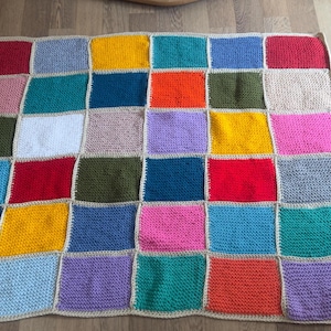 Könnte beinhalten: Eine handgefertigte Patchwork-Decke mit bunten Quadraten in Rot, Blau, Gelb, Grün und Rosa. Die Quadrate sind in einem Raster angeordnet und von einem neutralfarbenen Rand umgeben, was ein gemütliches Aussehen erzeugt.