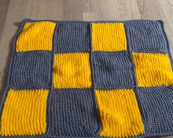 Könnte beinhalten: Handgestrickte Decke mit Schachbrettmuster. Die Decke hat abwechselnd gelbe und dunkelgraue Quadrate. Das Garn hat ein strukturiertes Aussehen, und die Decke ist sauber mit einem Rand versehen.