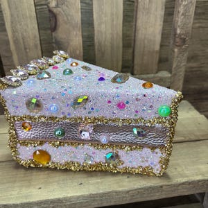 May include: A decorative, glitter-covered cake slice-shaped box. The box is white with iridescent glitter and adorned with colorful rhinestones and gold trim. The side of the box has a pinkish-gold band.