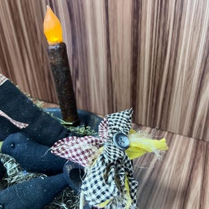 May include: A rustic centrepiece with a battery-operated candle with an orange flame. The arrangement includes black fabric crows and a fabric flower with a button, set in a metal bowl.