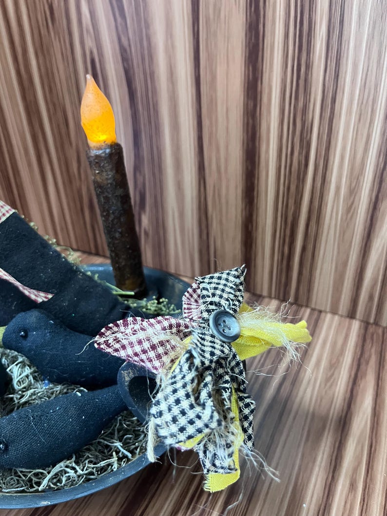 May include: A rustic centrepiece with a battery-operated candle with an orange flame. The arrangement includes black fabric crows and a fabric flower with a button, set in a metal bowl.