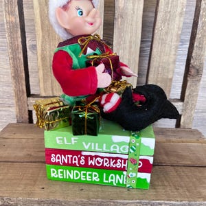 May include: A Christmas elf doll wearing a green and red outfit and a white-trimmed green hat. The elf is sitting on a stack of three painted wooden blocks that read "ELF VILLAGE", "SANTA'S WORKSHOP", and "REINDEER LANE". Several wrapped gifts are nearby.