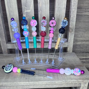 May include: A collection of beaded pens in various colors, including purple, teal, pink, and gold, displayed in a clear acrylic stand. Each pen features a decorative beaded top and a metal tip. Some pens are lying down.