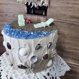 May include: A decorative cake with a beach theme. The cake features a miniature thatched-roof bar, beach chairs, a colorful ball, and blue crystal-like accents. Seashells adorn the sides, and the cake sits on a white lace doily.