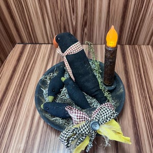 May include: A rustic arrangement featuring three black crow figures with red and white checked scarves, set in a bowl with faux greenery. A battery-operated candle with an orange flickering light and a decorative bow complete the display.