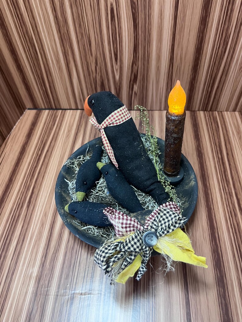 May include: A rustic arrangement featuring three black crow figures with red and white checked scarves, set in a bowl with faux greenery. A battery-operated candle with an orange flickering light and a decorative bow complete the display.