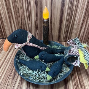 May include: A black bowl contains a black crow with an orange beak and a red and white checked ribbon. Three smaller crows with green beaks are in the bowl with a faux candle. The bowl is filled with decorative straw.