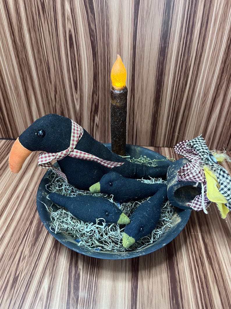 May include: A black bowl contains a black crow with an orange beak and a red and white checked ribbon. Three smaller crows with green beaks are in the bowl with a faux candle. The bowl is filled with decorative straw.