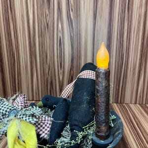 May include: A black bowl displays a decorative arrangement. It includes a black crow figure with a red and white checked scarf, a faux candle with an orange flickering light, and a yellow fabric accent. The arrangement is set against a wood-grain background.