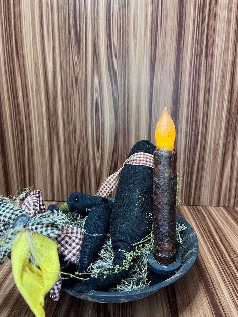 May include: A black bowl displays a decorative arrangement. It includes a black crow figure with a red and white checked scarf, a faux candle with an orange flickering light, and a yellow fabric accent. The arrangement is set against a wood-grain background.