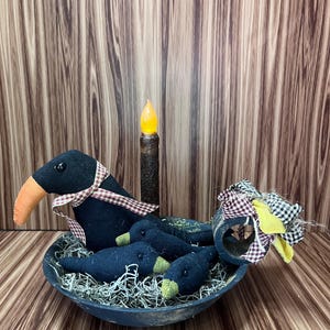 May include: A Halloween-themed decorative centrepiece. A black bowl holds three small crow figures and one larger crow figure. The crows have orange beaks and are in dried moss. A faux candle and fabric accents complete the arrangement.