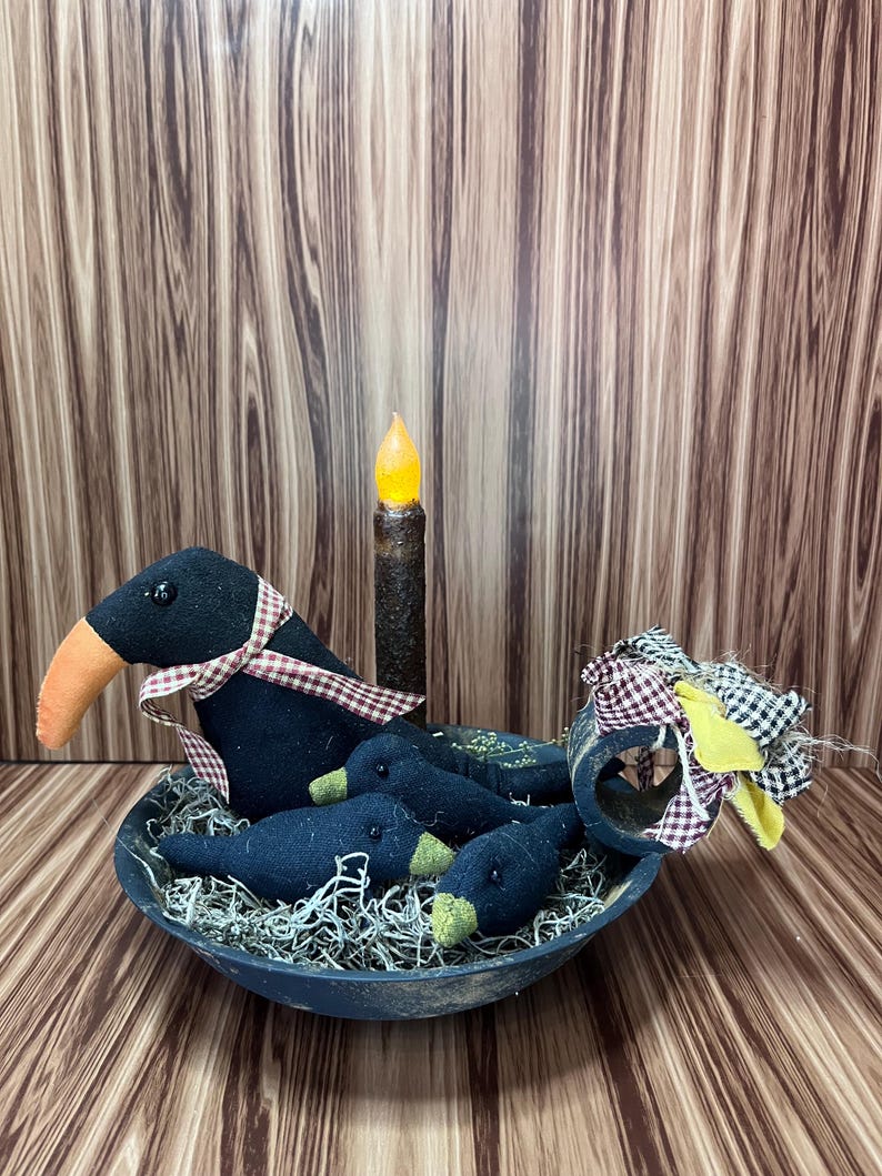 May include: A Halloween-themed decorative centrepiece. A black bowl holds three small crow figures and one larger crow figure. The crows have orange beaks and are in dried moss. A faux candle and fabric accents complete the arrangement.