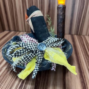 May include: A decorative arrangement in a black bowl. A black hat with a plaid scarf sits in the bowl. A bow made of yellow, black and white checkered, and plaid fabric is attached. A battery-operated candle with an orange flame is to the right of the hat.