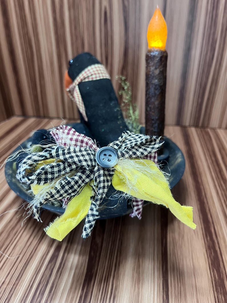 May include: A decorative arrangement in a black bowl. A black hat with a plaid scarf sits in the bowl. A bow made of yellow, black and white checkered, and plaid fabric is attached. A battery-operated candle with an orange flame is to the right of the hat.
