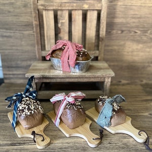 May include: Three loaves of bread wrapped in different colored checkered fabric bows, displayed on wooden cutting boards with handles. The loaves are on a wooden surface.