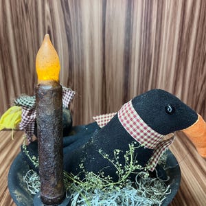 May include: A rustic arrangement featuring a black goose figurine with a red and white checkered scarf, nestled in a black bowl with faux greenery. A battery-operated candle with a flickering amber glow stands beside the goose.