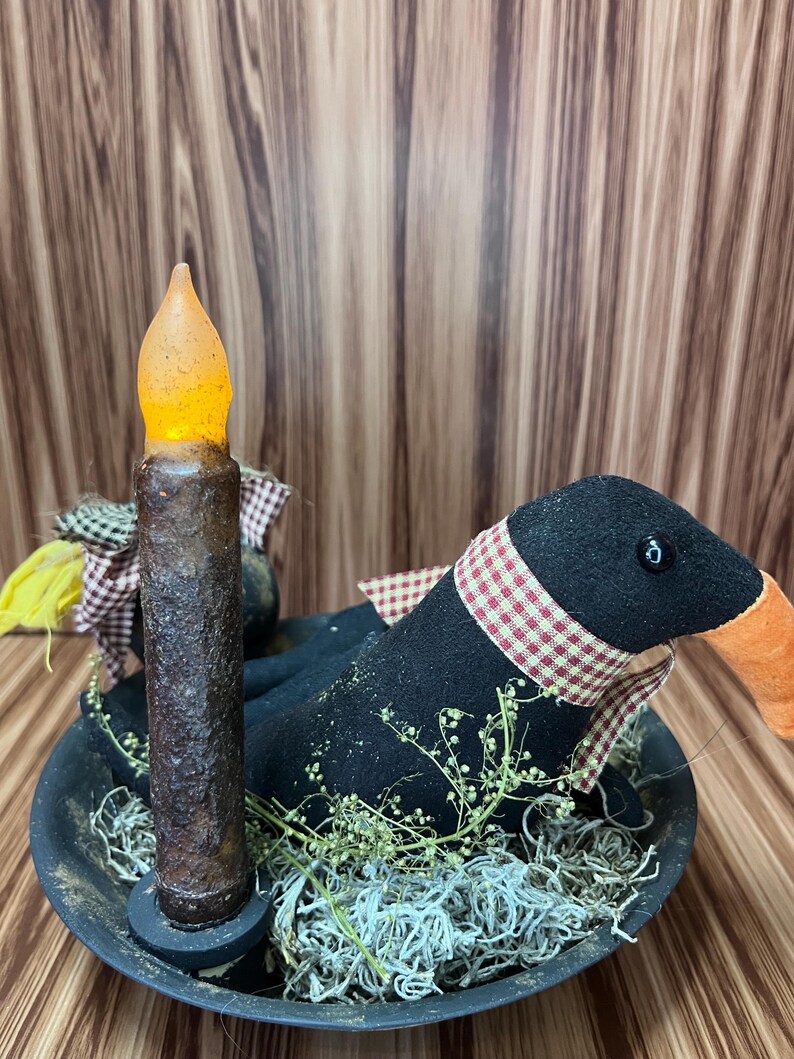 May include: A rustic arrangement featuring a black goose figurine with a red and white checkered scarf, nestled in a black bowl with faux greenery. A battery-operated candle with a flickering amber glow stands beside the goose.