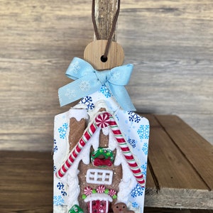 May include: A gingerbread house ornament with white frosting, red candy canes, and a green wreath. The ornament is decorated with blue snowflakes and a blue bow.