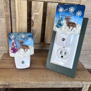 May include: Two white night lights with a winter scene on the front. The scene features a blue sky with snowflakes, a pine tree, and a deer. The larger night light is plugged into an outlet. The smaller night light is sitting on a wooden surface.