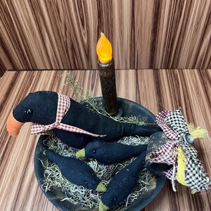 May include: A rustic black bowl holds a decorative arrangement featuring a black crow with an orange beak and a red and white checkered scarf, along with three smaller crow figures. A battery-operated candle and decorative accents complete the scene.