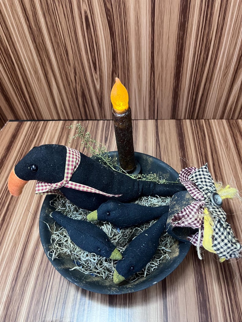 May include: A rustic black bowl holds a decorative arrangement featuring a black crow with an orange beak and a red and white checkered scarf, along with three smaller crow figures. A battery-operated candle and decorative accents complete the scene.