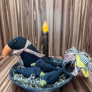 May include: A decorative arrangement featuring three black crow figures with orange beaks and plaid ribbon accents, nestled in a rustic blue bowl with faux moss. A battery-operated candle stands in the centre, creating a charming, country-themed display.