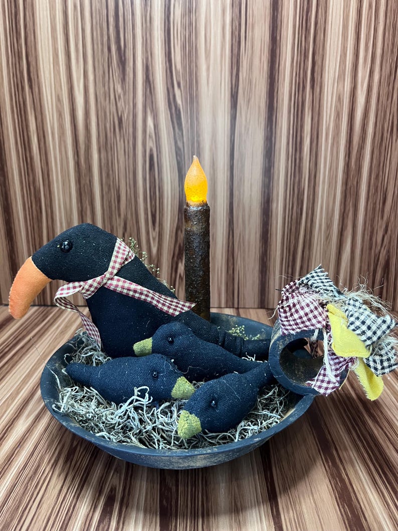 May include: A decorative arrangement featuring three black crow figures with orange beaks and plaid ribbon accents, nestled in a rustic blue bowl with faux moss. A battery-operated candle stands in the centre, creating a charming, country-themed display.
