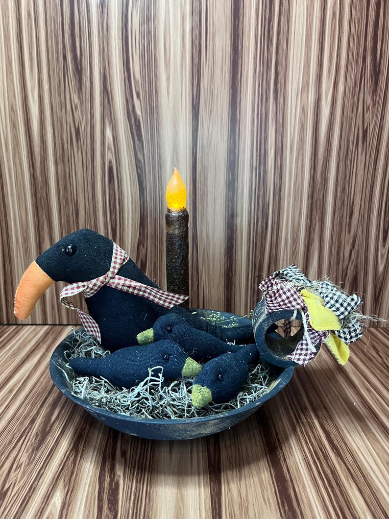 May include: A decorative arrangement featuring a black bowl filled with faux moss and several black bird figures with orange and green accents. A battery-operated candle and a decorative bow complete the rustic, farmhouse-style display.