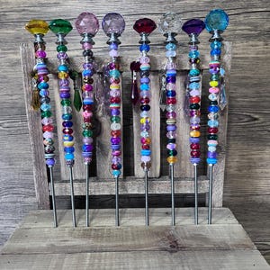 May include: A collection of eight decorative garden stakes. Each stake features a colorful beaded shaft and a faceted crystal or glass topper in various colors, including yellow, green, pink, purple, red, and blue. The stakes are displayed on a rustic wooden surface.