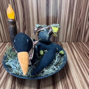 May include: A Halloween decoration featuring a black crow doll with an orange beak and a plaid scarf, in a black bowl with dried moss. A faux candle with an orange flame is also present. The arrangement is on a wooden surface.