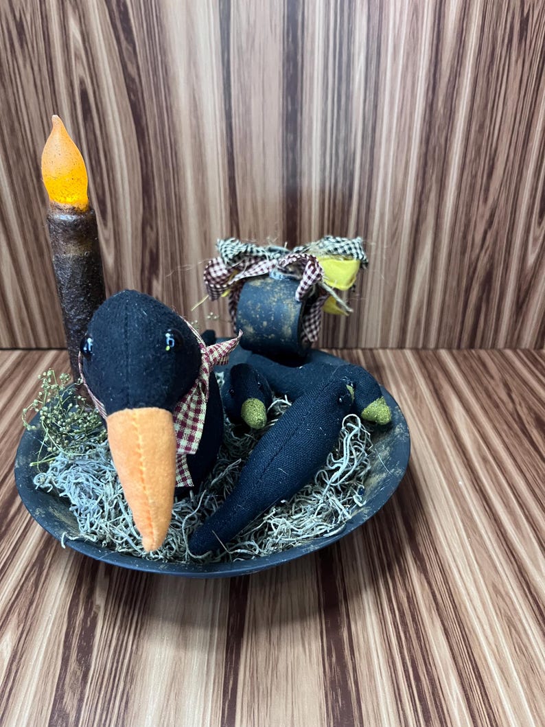 May include: A Halloween decoration featuring a black crow doll with an orange beak and a plaid scarf, in a black bowl with dried moss. A faux candle with an orange flame is also present. The arrangement is on a wooden surface.