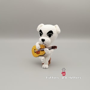 Puede incluir: Una figura de perro blanco de ganchillo tocando una guitarra amarilla y marrón. El perro tiene ojos negros y una nariz negra. La figura está sobre una superficie gris claro. El texto "Kittens and Mittens" está en la parte inferior.