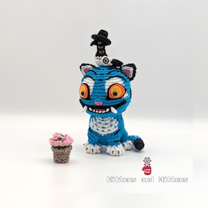 Pode incluir: Um gato azul de crochê feito à mão com detalhes brancos, olhos grandes cor de laranja e um pequeno chapéu preto. Uma pequena flor rosa em um vaso marrom fica ao lado do gato. O texto "Kittens and Mittens" está na parte inferior.