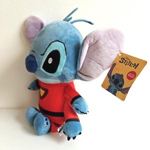 May include: A plush Stitch doll in a red jumpsuit with black accents. The blue alien character has large ears, a black nose, and a tag that reads "Disney Stitch". The doll is sitting on a white surface.