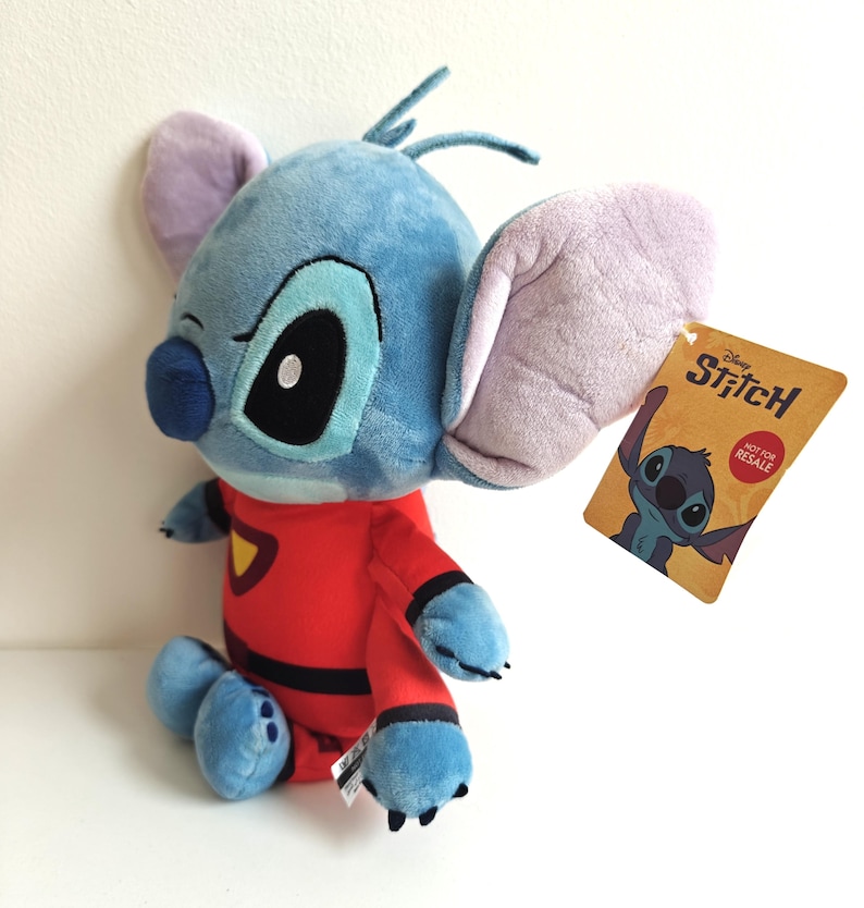 May include: A plush Stitch doll in a red jumpsuit with black accents. The blue alien character has large ears, a black nose, and a tag that reads "Disney Stitch". The doll is sitting on a white surface.