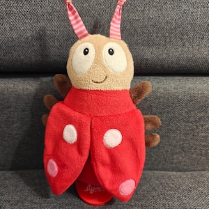 Puede incluir: Un juguete de mariquita de peluche con cuerpo rojo, lunares blancos y rosas y alas marrones. El juguete tiene una cabeza beige con ojos blancos grandes y antenas a rayas rosas y blancas. El juguete está sobre una superficie gris.