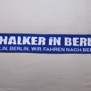 Może przedstawiać: Niebiesko-biały szalik z napisem "Schalker in Berlin" i logo "S04". Szalik ma również napis "Berlin, Berlin, wir fahren nach Berlin".