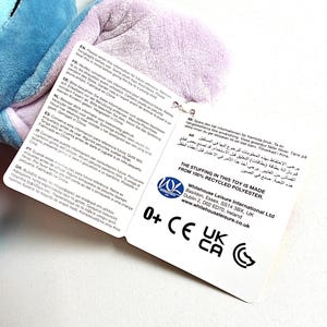 May include: A plush toy with blue and purple sections, with a tag attached. The tag includes text in multiple languages, the toy's stuffing is made from 100% recycled polyester, and the toy is suitable for ages 0+.