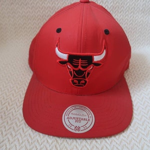 May include: Red adjustable baseball cap with a black and white Chicago Bulls logo. The cap has a white label that says "Adjustable Fit" and a black and white New Era logo.