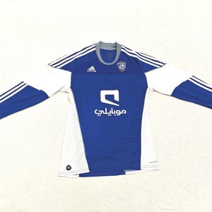 May include: A long-sleeved blue and white soccer jersey with the Adidas logo and three white stripes on the shoulders. The jersey has a crew neck and a team emblem on the chest. The word "موبايلي" is printed on the front.