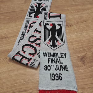 Könnte beinhalten: Hellgrauer Strickschal mit roten Fransen. Der Schal zeigt ein schwarzes Adleremblem und den Text "Wembley Final 30th June 1996". Das Wort "Deutschland" ist ebenfalls sichtbar. Wahrscheinlich ein Sport-Souvenir.