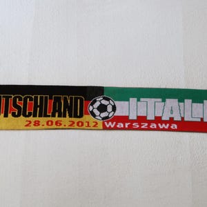 Puede incluir: Una bufanda tejida de color negro, amarillo, rojo y blanco con las palabras "DEUTSCHLAND" e "ITALIA" impresas en ella. La bufanda también presenta un balón de fútbol, el escudo de armas alemán y la bandera italiana. La fecha "28.06.2012" y la ciudad "Warszawa" también están impresas en la bufanda.
