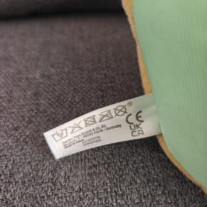 May include: A close-up of a plush toy with a green exterior and a yellow border. A white tag with washing instructions and the text "Simba Toys GmbH & Co. KG" is attached to the toy.