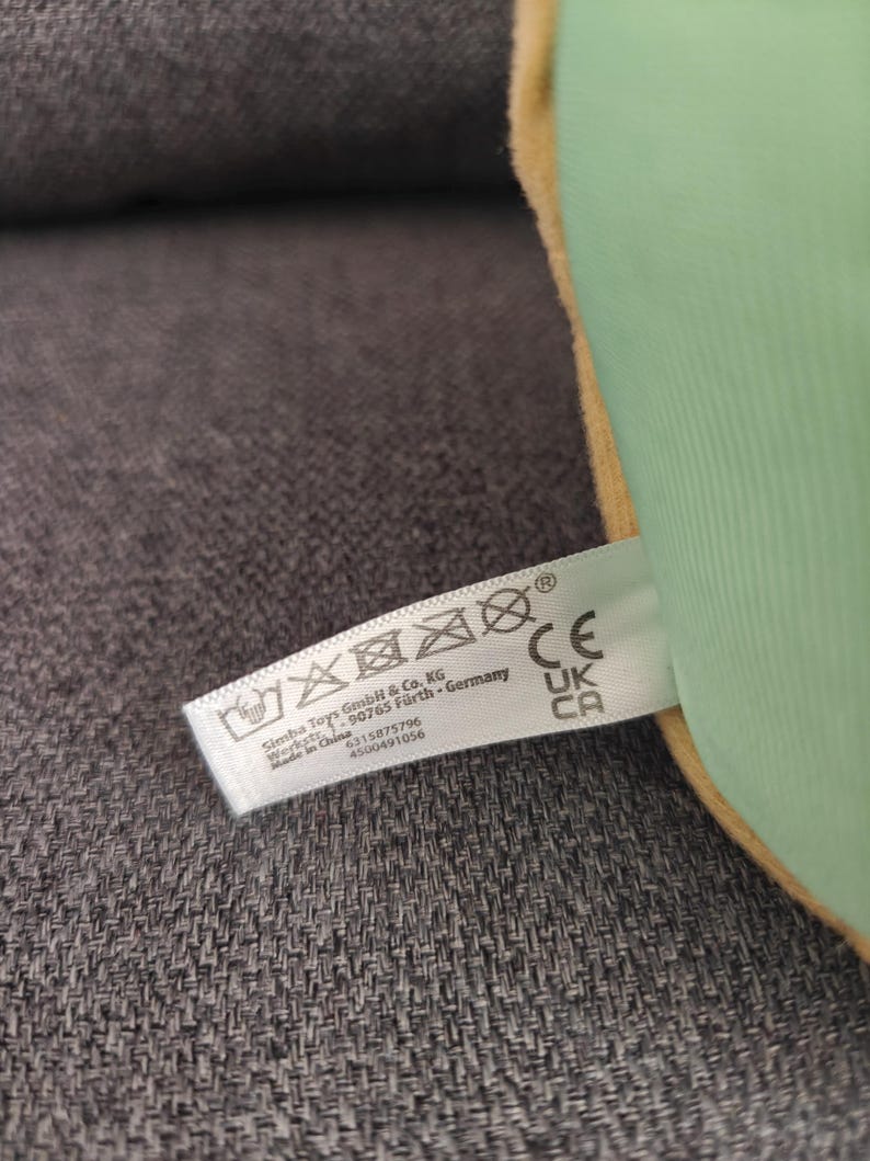 May include: A close-up of a plush toy with a green exterior and a yellow border. A white tag with washing instructions and the text "Simba Toys GmbH & Co. KG" is attached to the toy.