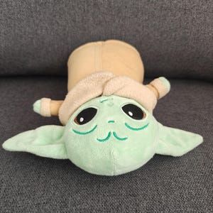May include: A plush toy of a character with large black eyes, green skin, and large ears. The character is wearing a tan coat and hood. The toy is lying on a gray surface.