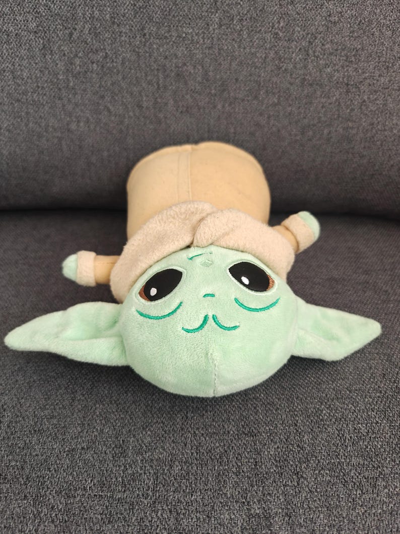 May include: A plush toy of a character with large black eyes, green skin, and large ears. The character is wearing a tan coat and hood. The toy is lying on a gray surface.