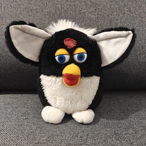 Puede incluir: Un juguete Furby en blanco y negro con orejas grandes, grises y blancas. Tiene ojos azules, un pico amarillo y una boca roja. Un parche rojo en forma de corazón con "I LOVE YOU" está en su frente. El juguete está sobre una superficie gris.