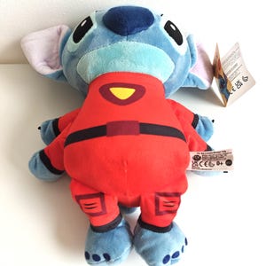 May include: A plush Stitch doll in a red jumpsuit with black accents. The doll has blue fur, large black eyes, and large ears. A yellow triangle is on the chest of the jumpsuit. A tag is attached to the doll.